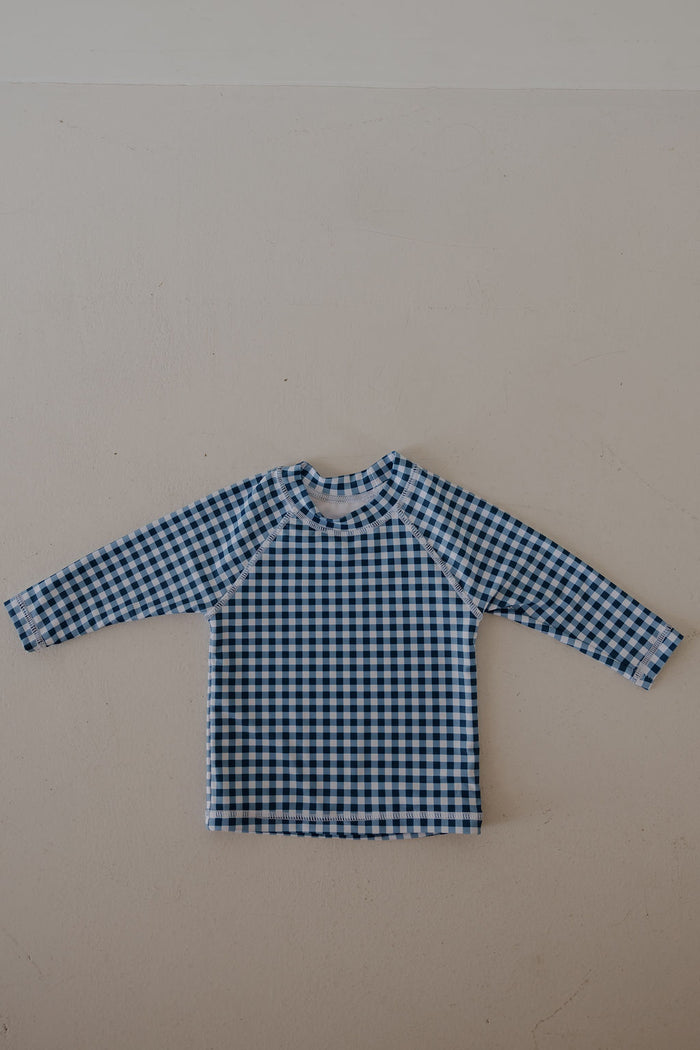 The forever french baby Child Rash Guard in blue gingham is laid flat on a light beige surface, making it a perfect summer option for kids.