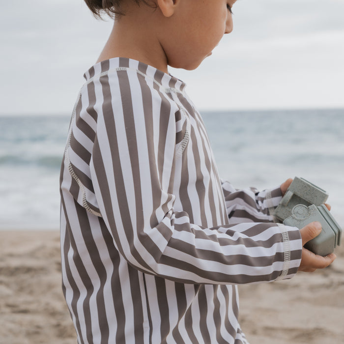A young child wearing the forever french baby Child Rash Guard in Olive Stripe stands on a sandy beach holding a small gray toy camera, with the ocean and sky behind them, ready for fun and sun protection.