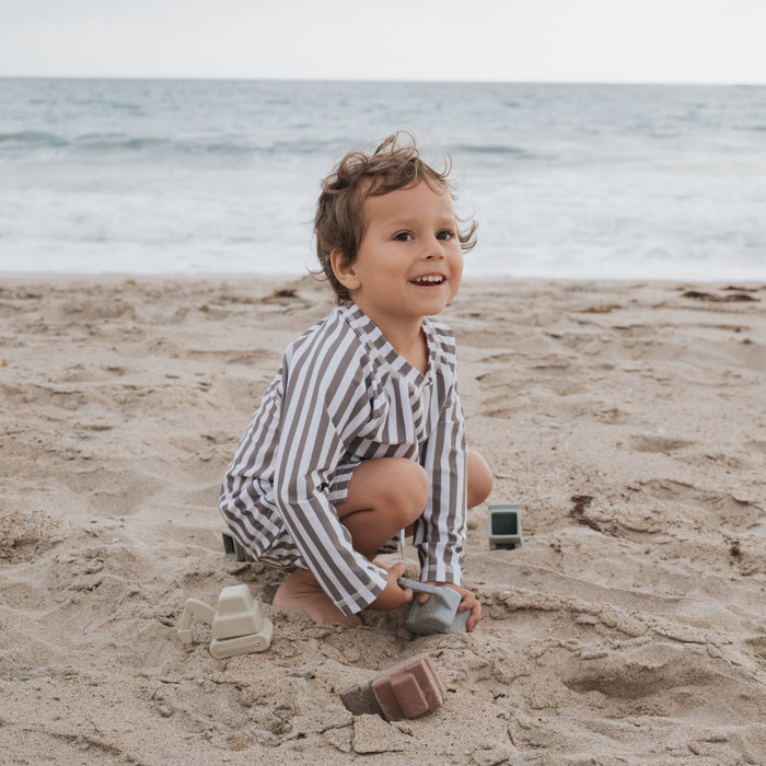 A young child wearing the forever french baby Child Rash Guard | Olive Stripe smiles and plays with toy blocks on a sandy beach, with ocean waves behind them. The rash guard offers sun protection for carefree play under a cloudy sky.