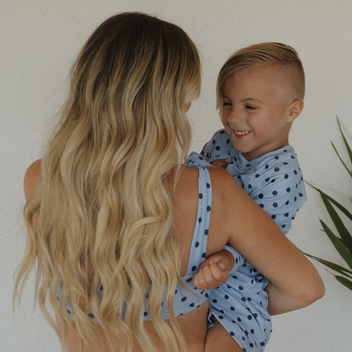 A woman with long, wavy blonde hair holds a smiling child wearing forever french baby’s Child Board Short | Sully. Both sport light blue family styles with dark blue polka dots, standing by a white wall and a green plant.