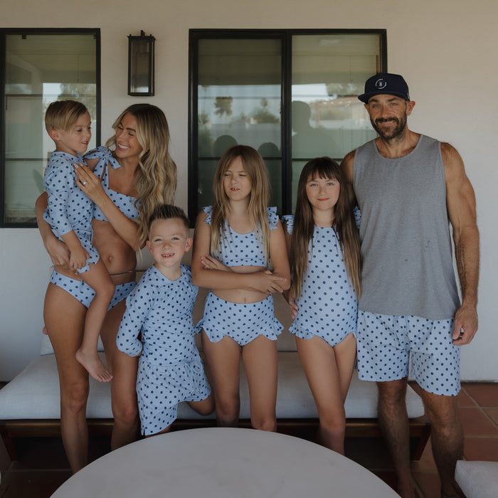 A smiling family of six stands outdoors in matching blue polka dot swimwear. The man wears forever french baby Men's Board Short | Sully and a tank top, while the rest wear coordinated polka dot swimsuits, two girls in the center, parents and two boys on each side.