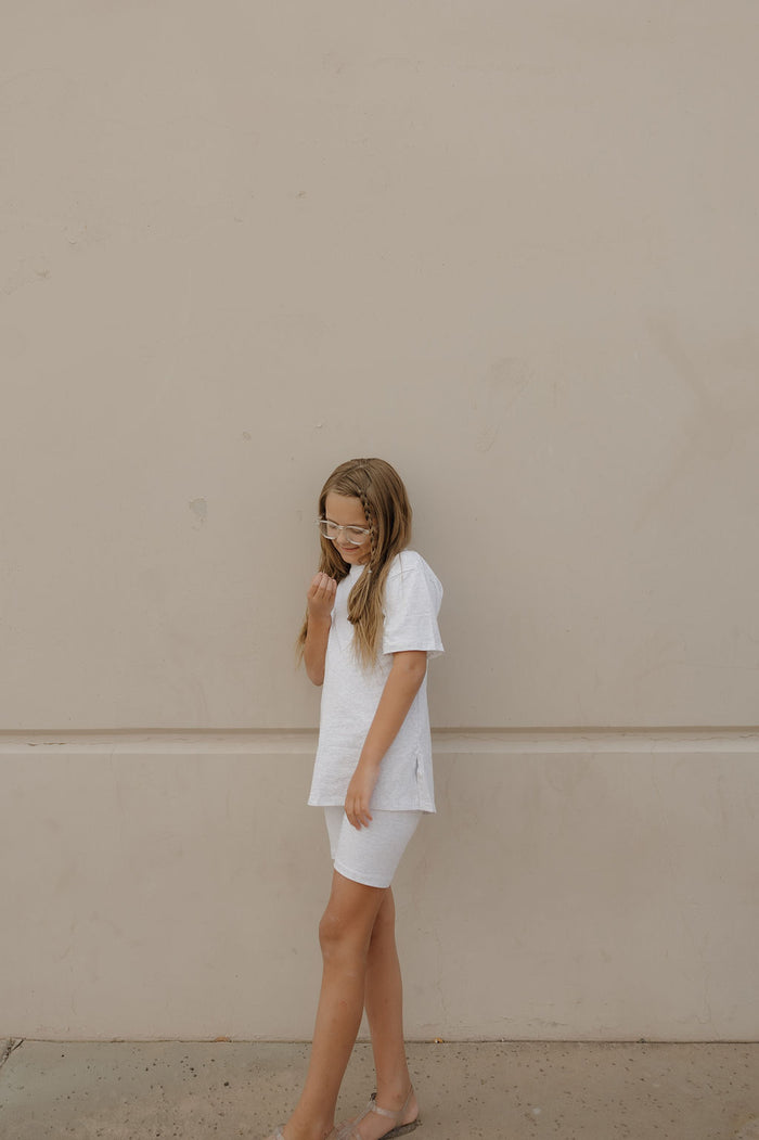 A young girl with long hair and glasses, wearing the forever french baby Child Short Set | Heather, stands against a plain beige wall, looking down thoughtfully.