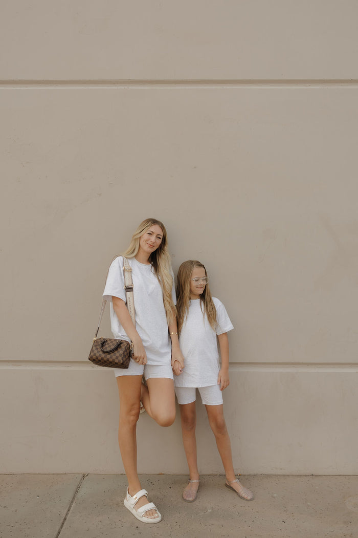 A woman and young girl stand against a beige wall, both in matching oversized tees and shorts. The girl wears the forever french baby Child Short Set | Heather. The woman has long blonde hair and carries a brown handbag. Both look relaxed with neutral expressions.