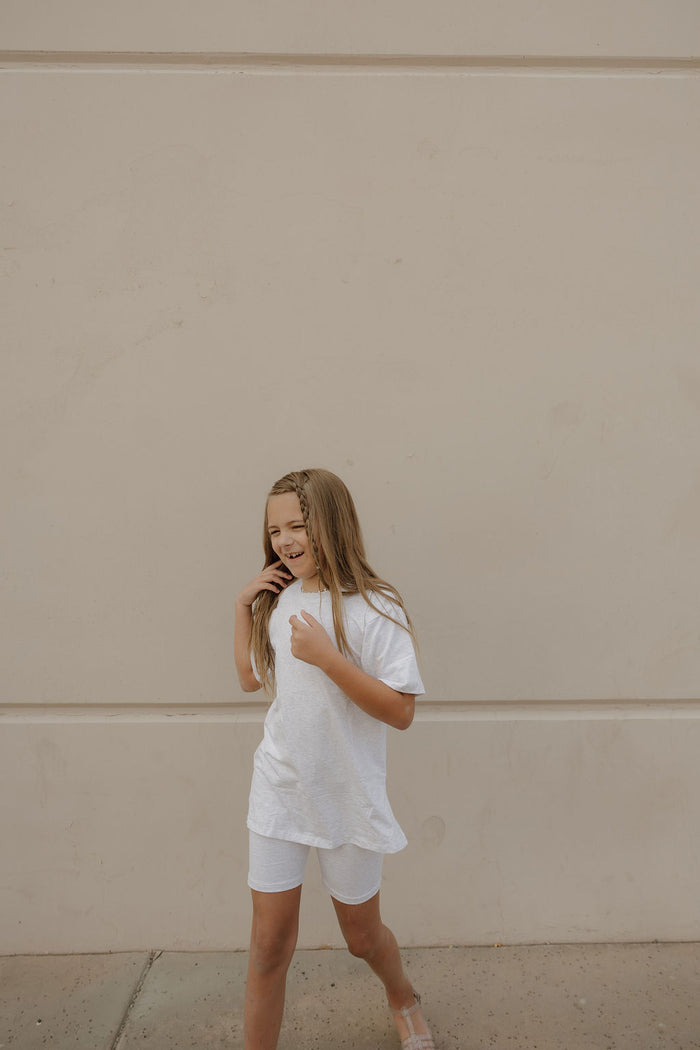 A young girl with long blonde hair wears the forever french baby Child Short Set | Heather as she smiles and looks to her left in front of a beige wall.