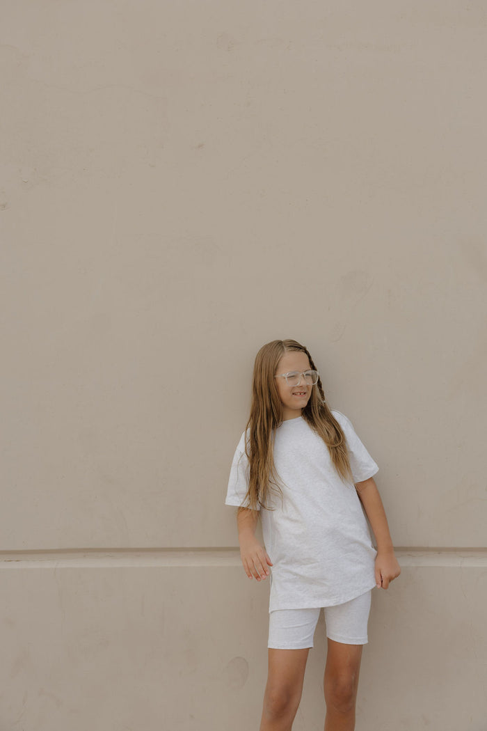 A young girl with long hair, wearing clear glasses and the forever french baby Child Short Set in Heather, stands against a plain beige wall, gazing slightly to the side.