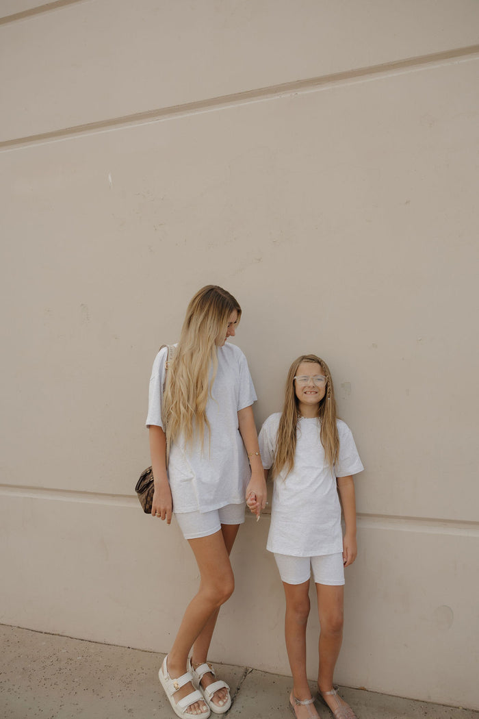 A woman and a young girl stand against a beige wall, smiling and holding hands in matching forever french baby Women's Short Set | Heather outfits with oversized tees. The woman has long blonde hair and sandals; the girl wears glasses.