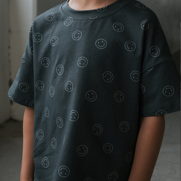A child models the forever french baby Child Short Set | Smiley, showing a dark gray cotton blend T-shirt with small white smiley faces. The indoor photo is cropped to highlight the upper body, with the head partially out of frame.