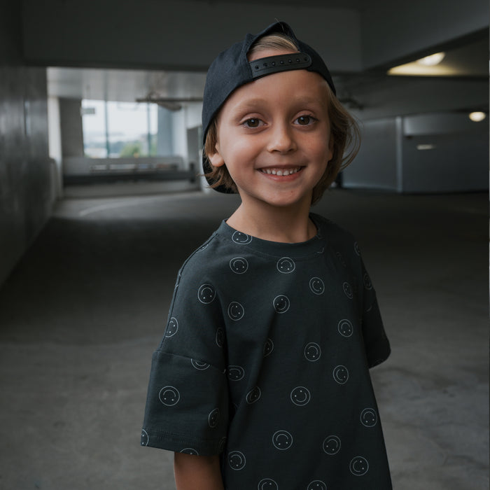 A young boy smiles in a dimly lit parking garage wearing the forever french baby Child Short Set | Smiley, featuring a dark cotton-blend top with smiley face patterns and a backward black cap.