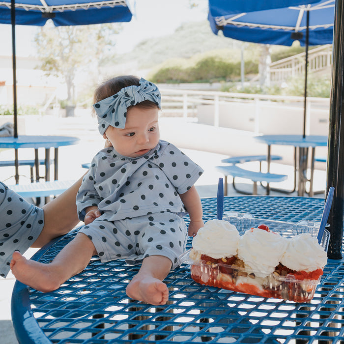 A baby wearing the forever french baby "Child Short Set | Sully" sits on a blue metal table, gazing at a tray of strawberry shortcake with whipped cream on a sunny patio.