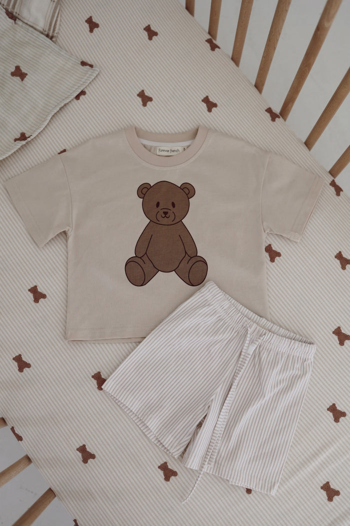 The forever french baby Child Short Set | Teddy features a beige toddler tee with a brown bear print and light striped shorts, displayed on a cream crib sheet with small brown bear faces.