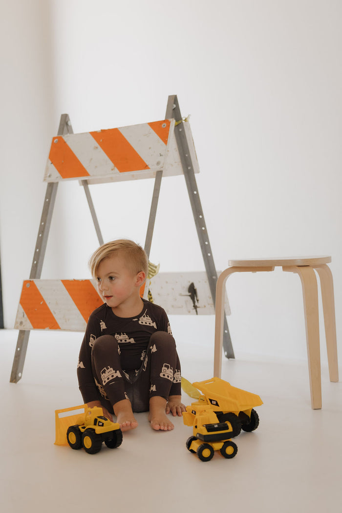 A young child in the forever french baby Child Sleepy Time Set | Bulldozer sits barefoot on a white floor, surrounded by yellow toy trucks, with orange and white construction barriers and a light wooden stool behind them.