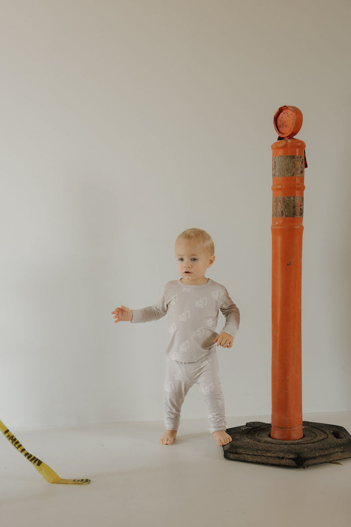 A toddler in the forever french baby Child Sleepy Time Set | Excavator stands indoors beside a tall orange traffic post on a white floor, with part of a yellow and black striped object visible on the left.