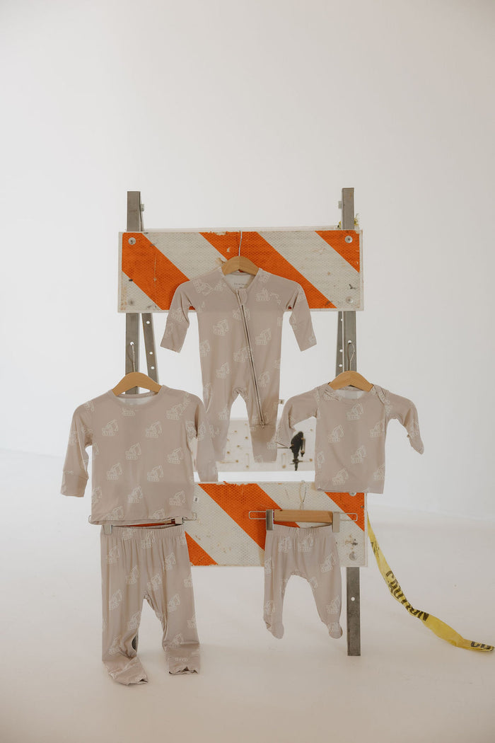 Four hypoallergenic, light beige pieces from the forever french baby Child Sleepy Time Set | Excavator, featuring a white animal print, hang on an orange-striped construction barricade in a white studio. A yellow caution tape is seen on the right.