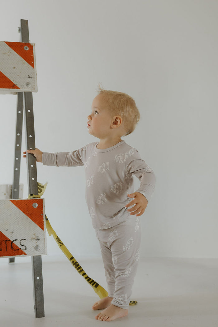 A barefoot toddler wears the forever french baby Child Sleepy Time Set | Excavator, standing indoors beside a metal traffic barricade with red and white stripes and yellow caution tape, looking up and to the side.