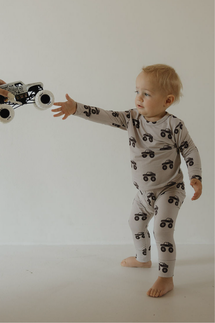 A toddler in the forever french baby Bamboo Footless Sleepy Time Set | Monster Truck reaches out for a toy monster truck being handed to them against a plain, light background.