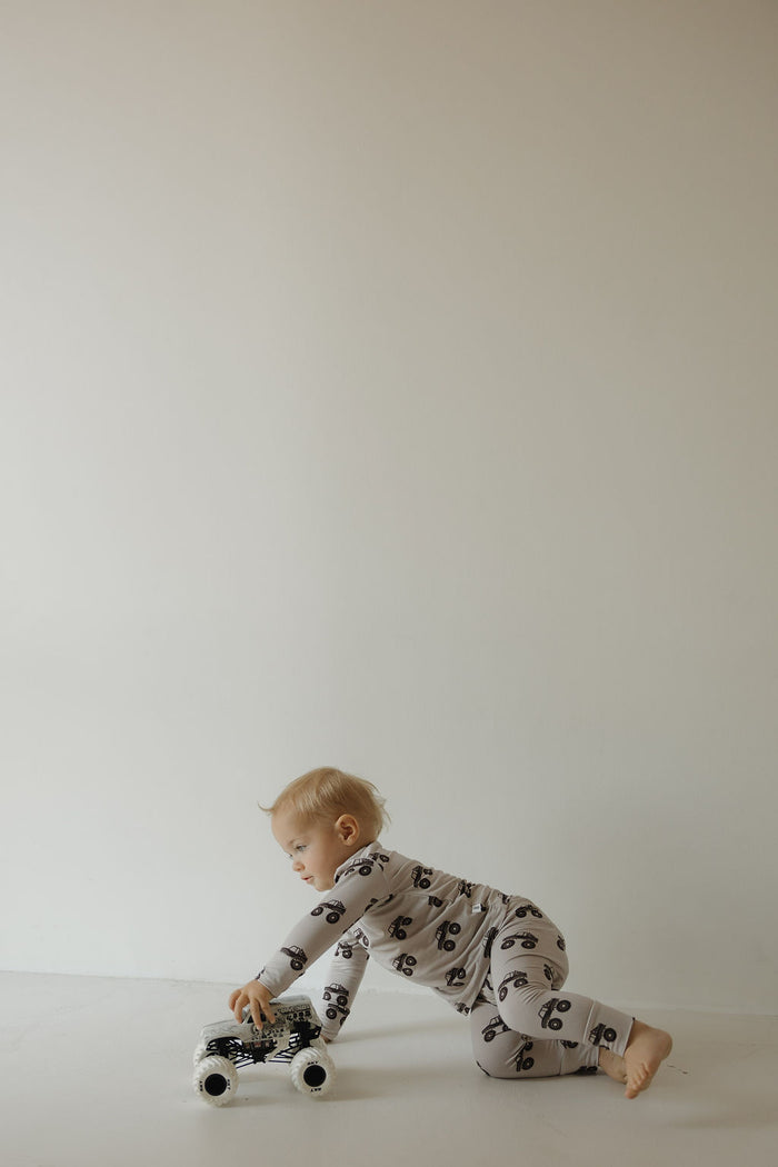 A young child in the forever french baby Bamboo Footless Sleepy Time Set | Monster Truck crawls and plays with a black and white toy car on a plain white background.