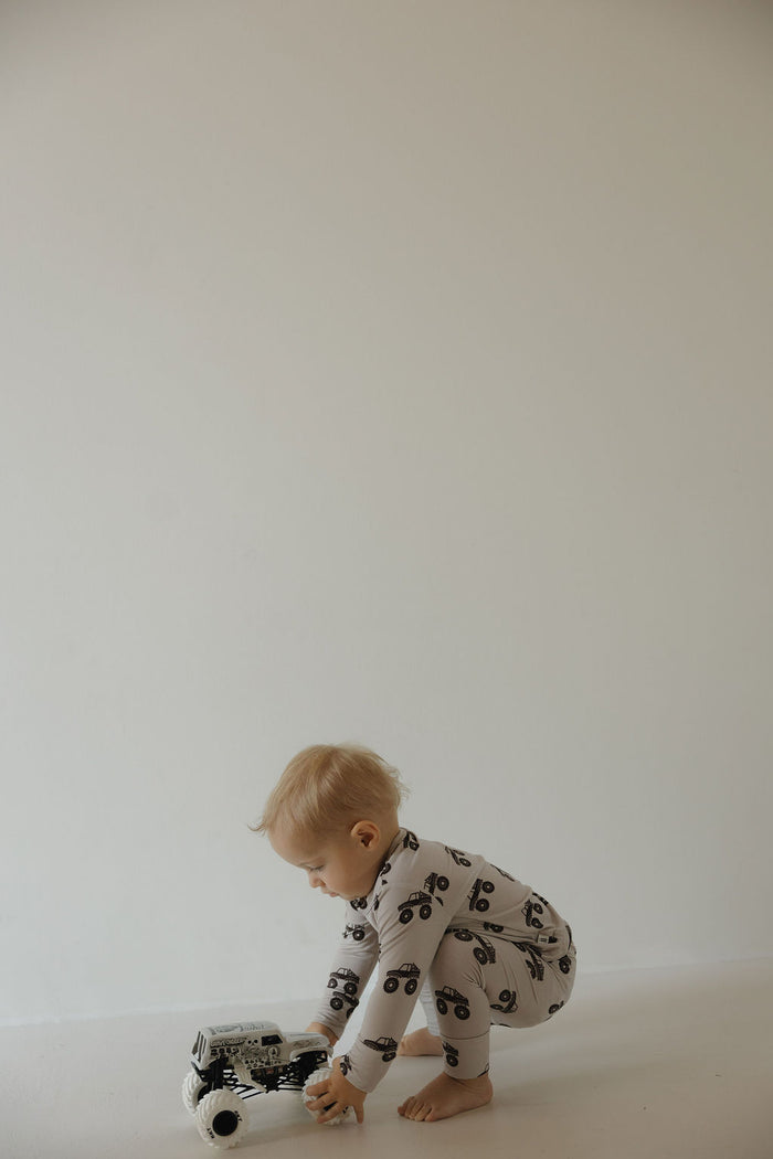 A young child in the forever french baby Bamboo Footless Sleepy Time Set | Monster Truck crouches on the floor, playing with a toy truck against a plain, light-colored background.