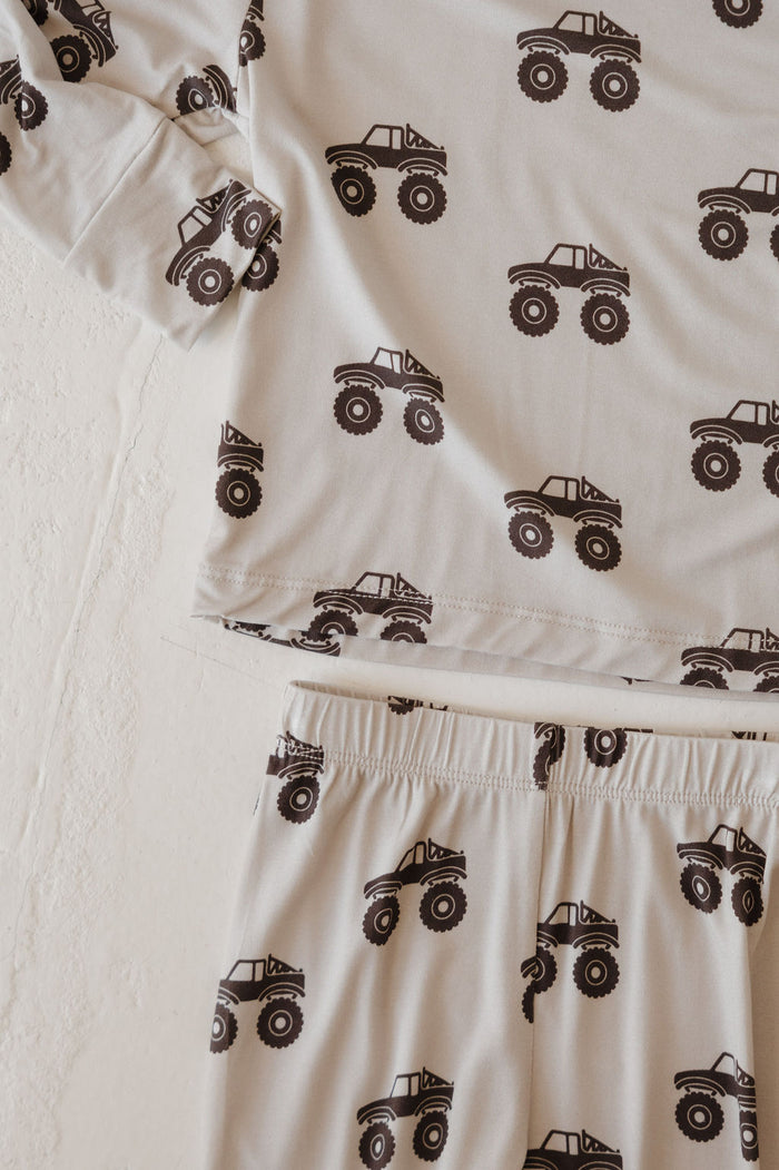 A close-up of the forever french baby Bamboo Footless Sleepy Time Set | Monster Truck, a hypo-allergenic, cream pajama set with black monster truck prints on the long-sleeve top and pants, laid out on a textured surface.