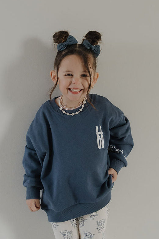 A young girl with brown hair in two buns and blue bows smiles at the camera, wearing the forever french baby Child Sweatshirt | ff Blue with a white "f", and white patterned pants, standing against a plain light-colored wall.