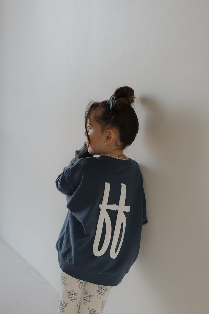 A young child with a bun stands against a white wall, wearing the forever french baby Child Sweatshirt | ff Blue with large white "ff" letters on the back and light floral print pants.