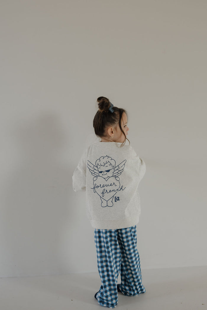 A young child with brown hair in a bun wears the forever french baby Child Sweatshirt | ff Los Angeles, featuring an angel graphic and "forever friend" on the back, paired with blue checkered pants against a plain light background.