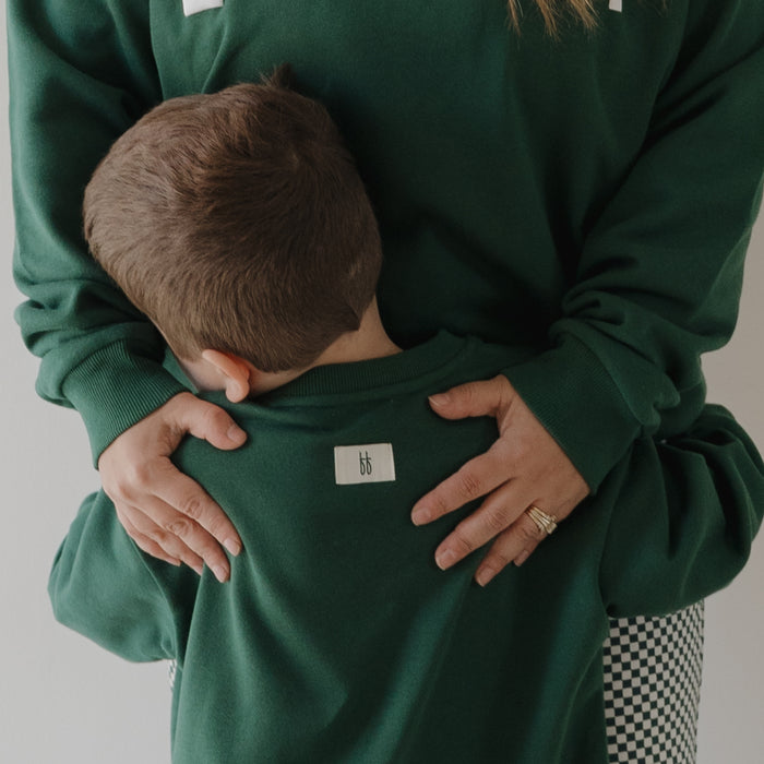 A child with short brown hair, wearing the forever french baby Child Sweatshirt | Lucky, hugs an adult in a matching green sweatshirt. Their torsos are shown—perfect for coordinating family outfits.