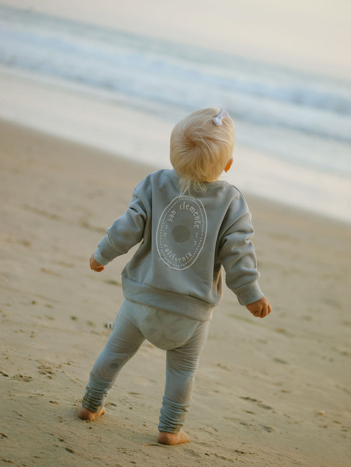 A young child in the forever french baby Child Sweatshirt | San Clemente stands barefoot on the sandy beach, gazing at ocean waves under a soft sky—a perfect coastal keepsake moment.