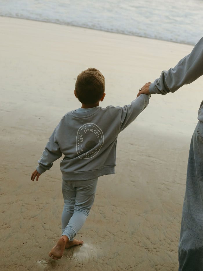 A young child wearing the forever french baby Child Sweatshirt | San Clemente in light gray walks barefoot on the beach, holding an adult's hand. The ocean and wet sand are visible in the background, with the adult partially out of frame.