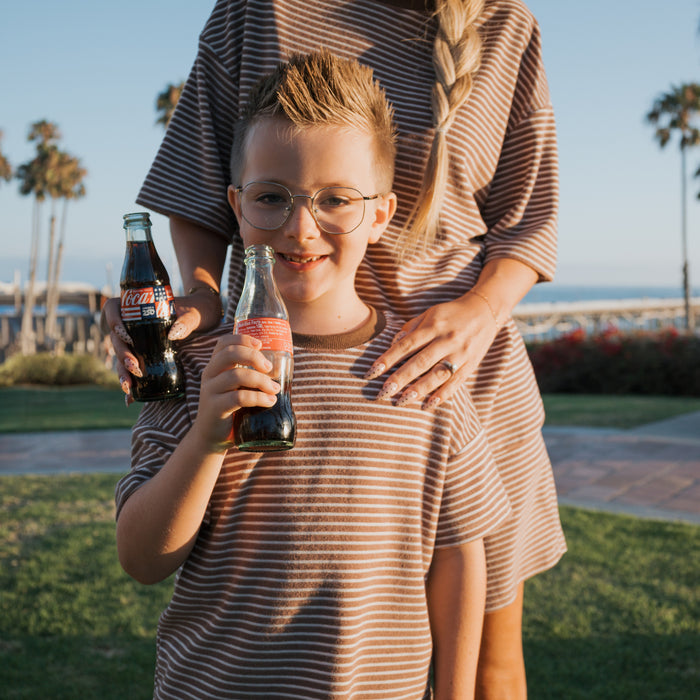 A smiling boy wears the forever french baby Child Terry Short Set | Cola Stripe, holding a Coke bottle beside a woman in a matching dress. They stand outdoors with palm trees, showing off their coordinated family look.
