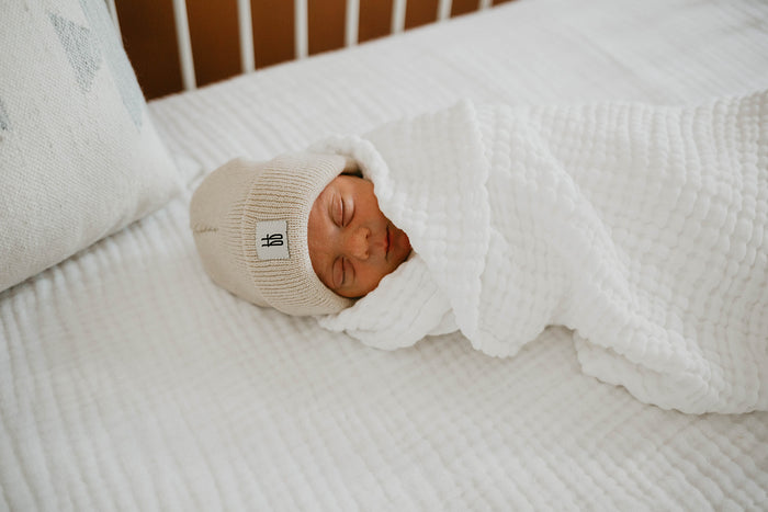 A newborn baby sleeps peacefully, swaddled in a soft white blanket and wearing a beige knitted hat adorned with a small Forever French Baby logo. The baby is lying on a "Crib Sheet | Cloud" by forever french baby, with a pillow nearby, creating a serene and cozy scene. Remember to wash with like colors and tumble dry on low.