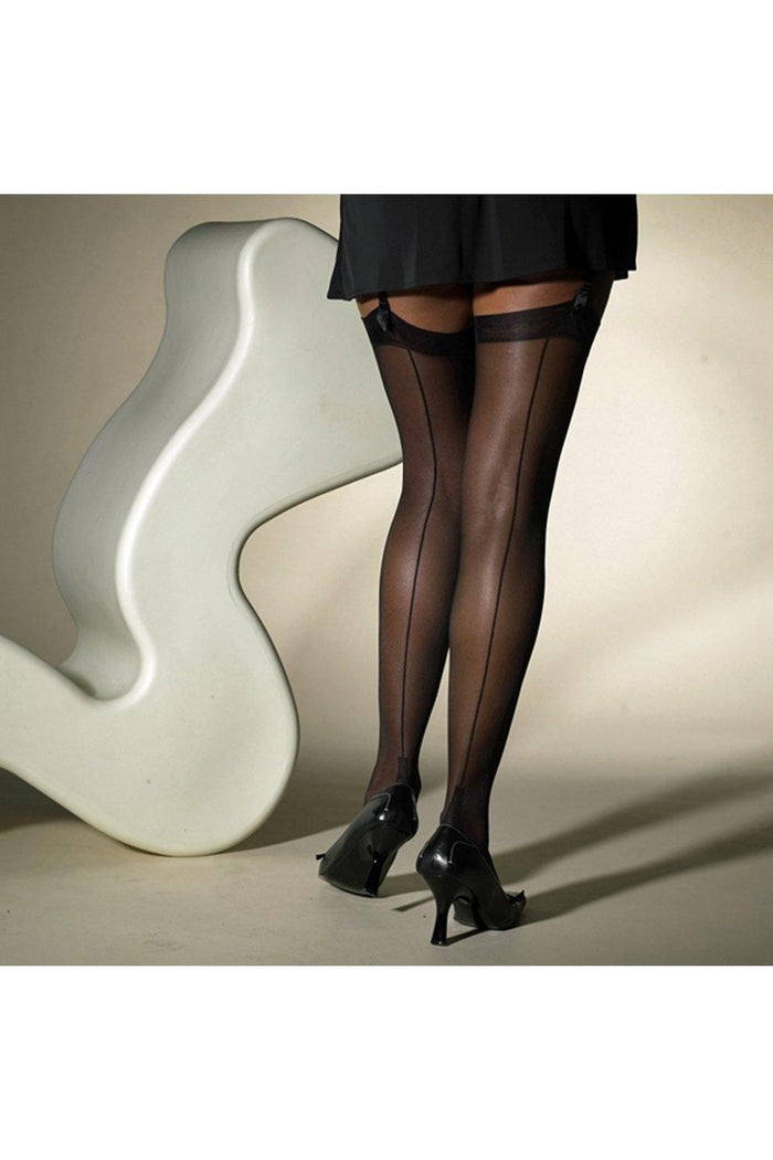 Model wearing black sheer seamed  stockings and high heels in front of a white chair.
