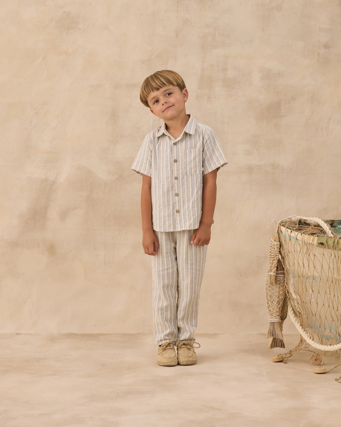 A young boy with light brown hair stands on a beige floor wearing the Rylee + Cru Ethan Trouser in Nautical Stripe, paired with a matching top. He leans to one side, with a basket and textured wall in the background.