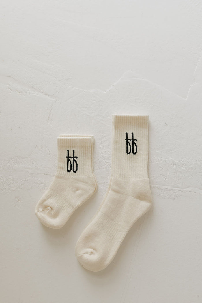 Two "ff Crew Socks | Vanilla" by forever french baby rest on a textured off-white surface. These cream-colored crew socks, ideal for daily wear, feature small black letters on the cuffs. One sock is larger, indicating they form a matching set for different sizes.