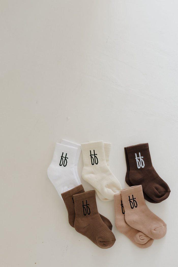 A set of six ff Crew Socks | Cocoa by forever french baby sits on a light surface, featuring shades of brown and cream. Each family-friendly pair displays a small black logo with two stylized "b" letters, capturing the brand's charm.