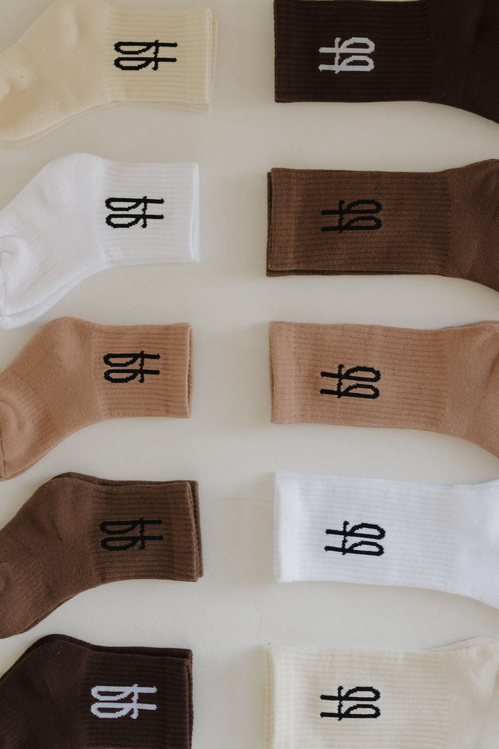 Forever French Baby's "ff Crew Socks | Cocoa" are displayed in rows on a flat surface, featuring various shades of brown, beige, and white. Each sock displays a black emblem near the cuff.
