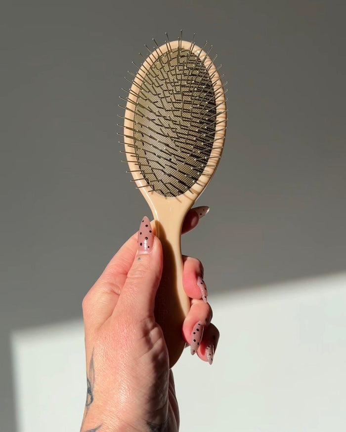 A hand with pink, black-speckled nails holds up the forever french baby ff Hair Brush, a beige detangling brush with stainless steel bristles, against a light gray background.