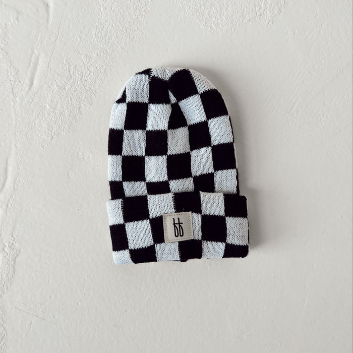 A black and white checkerboard-patterned ff Knit Beanie from forever french baby, featuring a small rectangular logo tag with stylized initials "ff" at the front. Made from breathable yarn, the hat is laid flat against a textured off-white background.