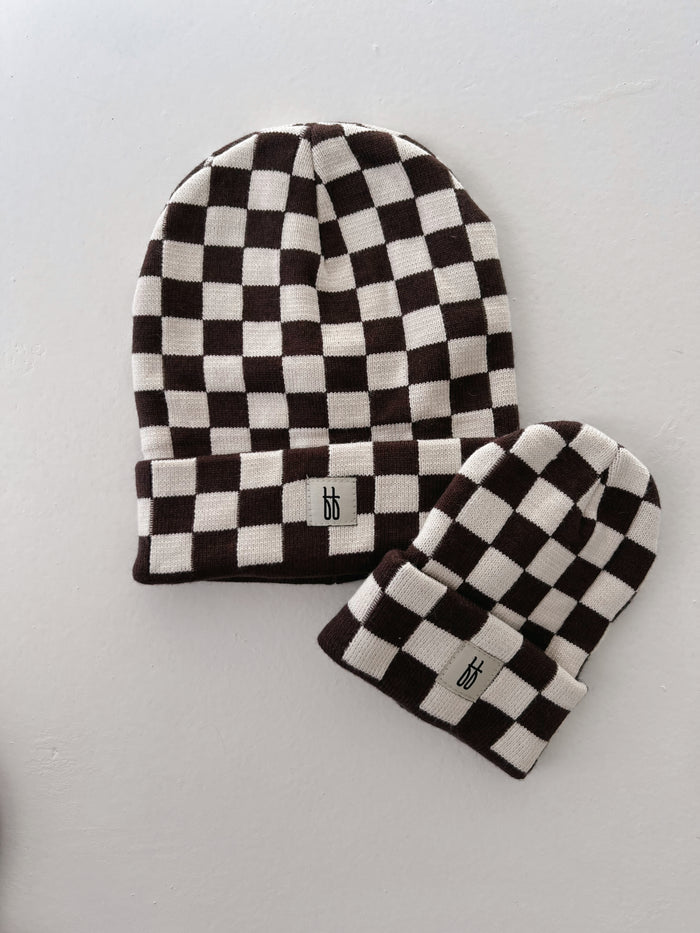 Two Coffee & Cream Checkerboard knit beanies from Forever French Baby are displayed against a white background. The larger beanie has a folded brim showcasing its classic ribbed design, while the smaller one is more narrow. Both feature a black and white checkered pattern and a small tag with the letters "bb" on the front.