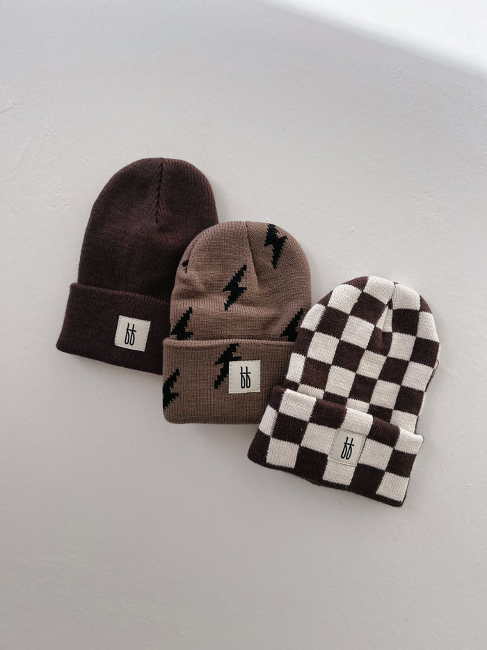 Three beanies are displayed on a white surface. The first is a plain, dark brown knit beanie. The second is light brown with a black thunderbolt pattern. The third, the "ff Knit Beanie | Coffee & Cream Checkerboard" from Forever French Baby, has a black and white checkered design. Each beanie features a small white logo patch with the letters "bb.