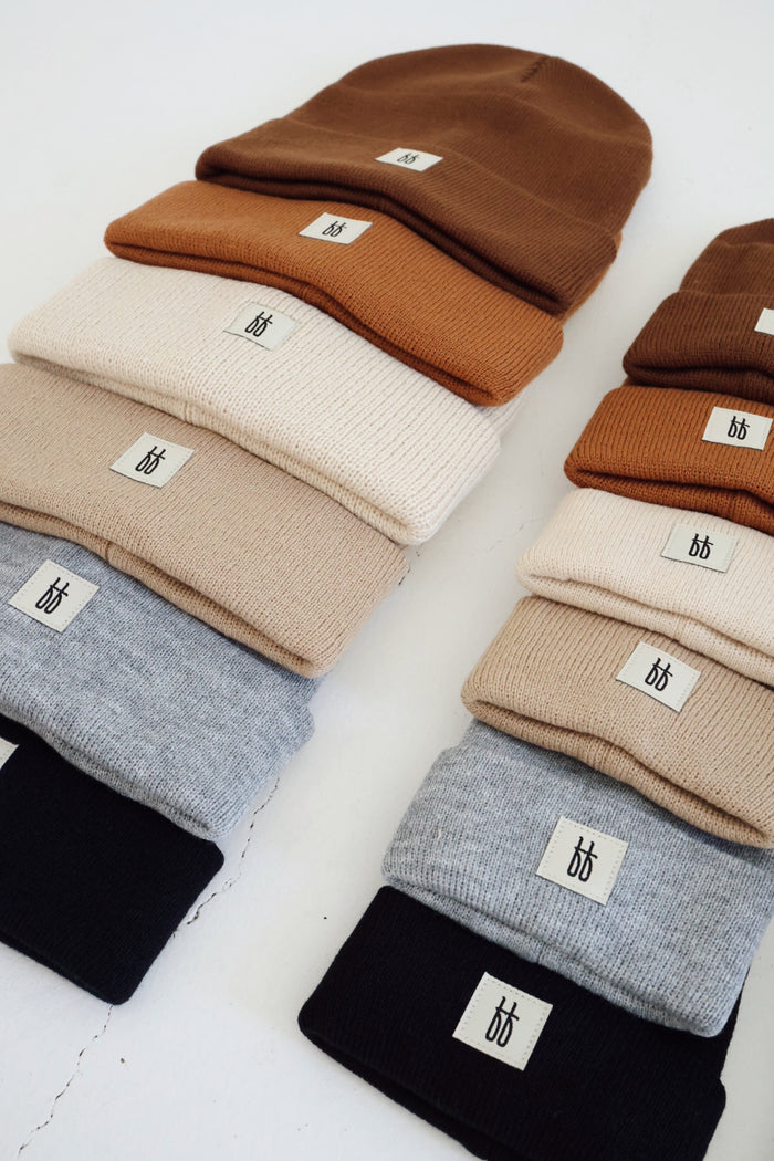 A collection of beanies, including the ff Knit Beanie | Adult from forever french baby, is neatly arranged in four stacks on a white surface. These beanies come in various earthy tones like brown, beige, light gray, and black. Each ribbed design features a small white rectangular tag with a minimalist logo design.