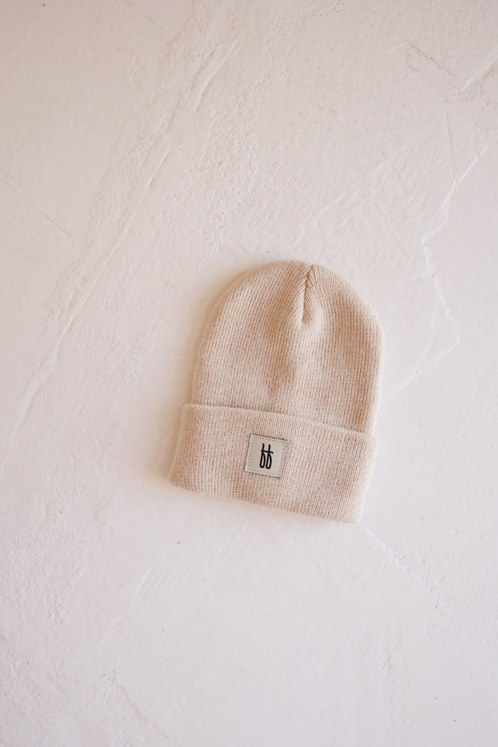 An oatmeal-toned ff Knit Beanie with a folded brim and ribbed design rests on a textured white surface. The small rectangular tag on the brim features Forever French Baby's minimalist, stylized "bb" logo in black.