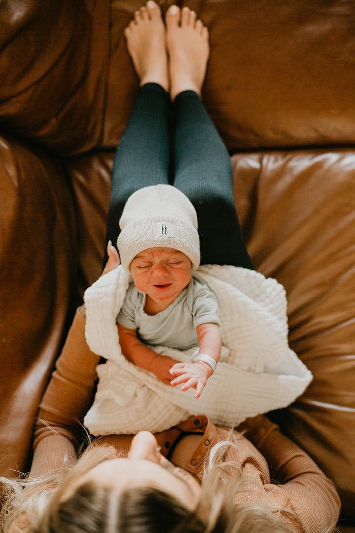 A person is sitting on a brown leather couch with their legs stretched out, holding a crying newborn baby wrapped in a white blanket. The baby, dressed in a light-colored outfit and wearing an adorable ff Knit Beanie | Oatmeal from forever french baby, snuggles close for comfort.