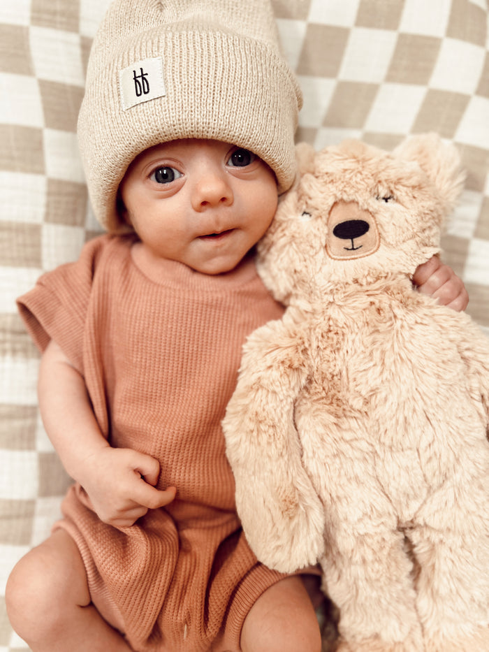 A baby wearing a Forever French Baby ff Knit Beanie in Oatmeal and a brown outfit is sitting on a checkered blanket. The baby is holding a fluffy teddy bear and looking at the camera with wide eyes, embodying the charm of Forever French Baby fashion.