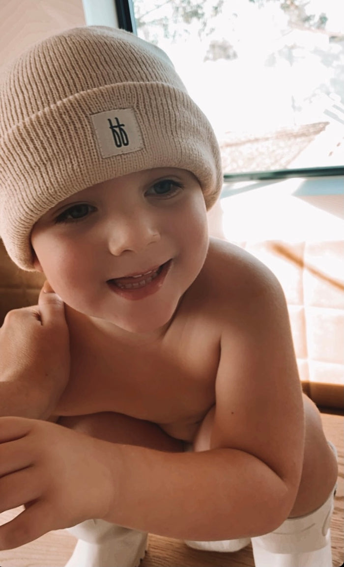 A young child smiling, wearing an oatmeal-colored ff Knit Beanie with a ribbed design, bare shoulders, and visible arms, crouched near a sunlit window. The beanie features a small logo on the front from forever french baby.