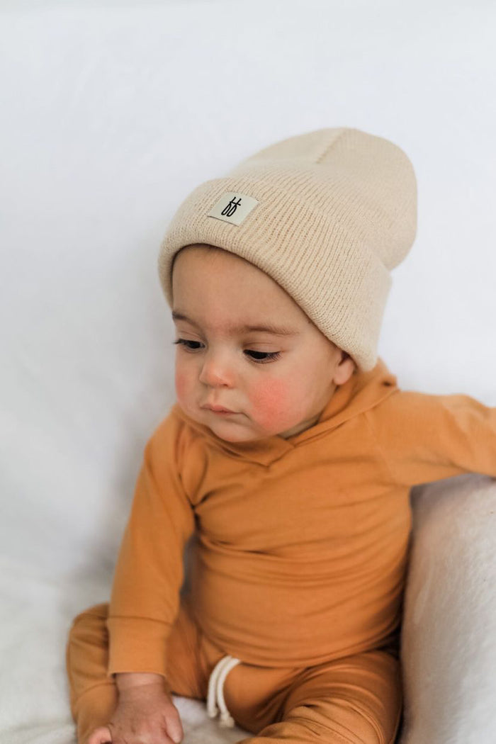 A baby dressed in the ff Knit Beanie | Oatmeal and an orange outfit sits on a beige blanket. The baby gazes downward with a calm expression against a white background, embodying the timeless elegance of forever french baby.