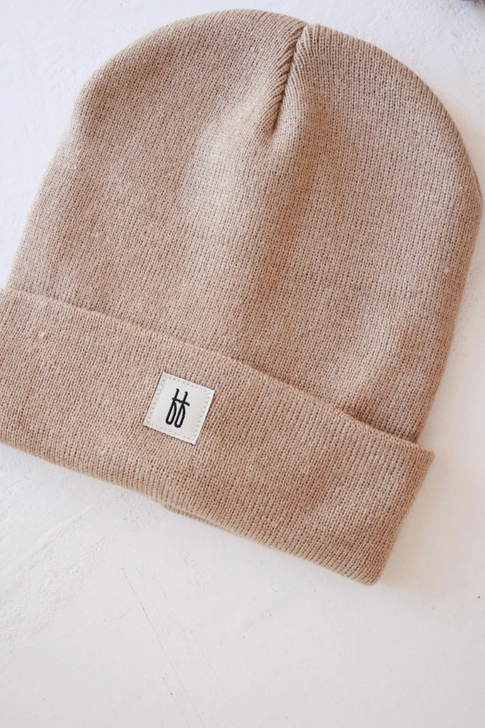 A ff Knit Beanie in tan by forever french baby is placed on a white surface. The beanie features a small white rectangular tag with a minimalist black logo sewn onto its folded brim, showcasing the ribbed design. The knit texture and breathable yarn of the ff Knit Beanie are clearly visible.