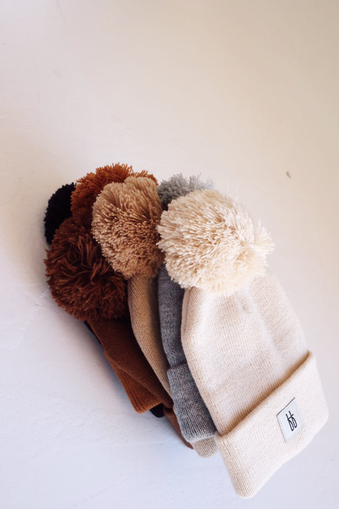 A stack of forever french baby ff Knit Pom Top Beanies, each topped with a pom-pom and featuring a classic ribbed design, is arranged against a white background. The colors include cream, gray, brown, and black. The beanies have a small, stylized forever french baby logo tag near the cuff.