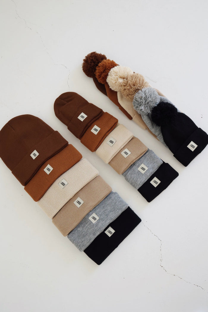 An arrangement of the "ff Knit Beanie | Adult" from forever french baby is displayed in three vertical columns, showcasing various shades of brown, beige, gray, and black. Each breathable yarn beanie features a small white tag with a simple emblem. The right column includes beanies adorned with pom-poms on top. The background is a light-colored surface.