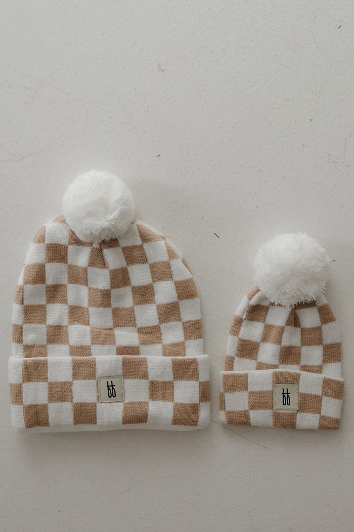 Two ff Knit Pom Top Beanies in Tan Checkerboard by forever french baby—one adult and one baby size—are side by side on a light surface, each with a "bb" logo label on the folded brim and a white pom-pom on top.