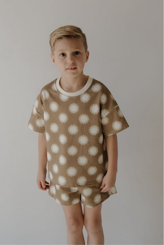 A young child with short blonde hair wears the French Terry Short Set | Sol by forever french baby, featuring a soft short sleeve top and matching shorts with a white sunburst pattern, standing against a plain background and smiling slightly at the camera.